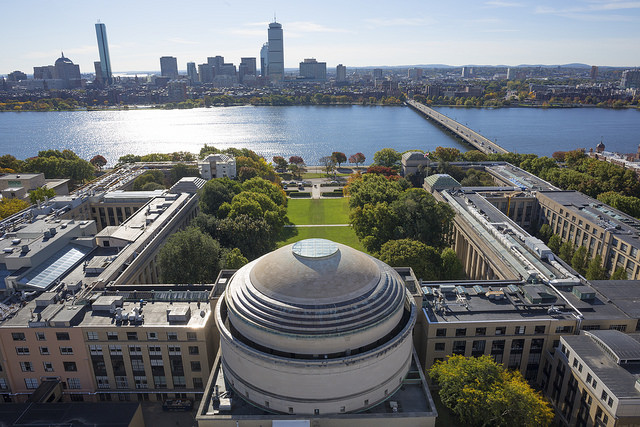 Aerial photo of the MIT dome and other nearby buildings, with the Charles River and Boston skyline in the background.