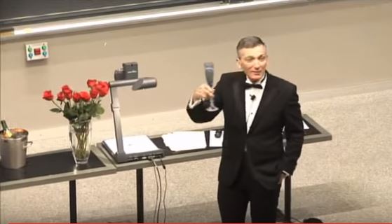 Screenshot of video with tuxedo-clad professor at the front of lecture hall, holding up a glass of champagne.