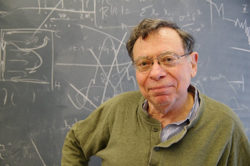 Photo of a smiling man in front of a chalkboard covered in math and diagrams.