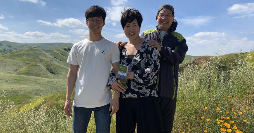 Photo of Kyle with his parents, outdoors on a grassy hillside.